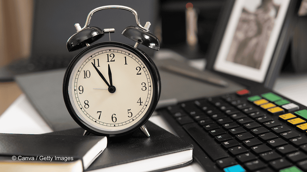 Auf einem Schreibtisch liegen Notizbücher und eine Tastatur. Auf den Notizbüchern steht ein schwarzer analoger Wecker, der symbolisch für die Arbeitszeit stehen soll. Im Hintergrund steht ein gerahmtes Foto, was allerdings nur verschwommen zu sehen ist. Im Text geht es um Arbeitszeitverlängerung.