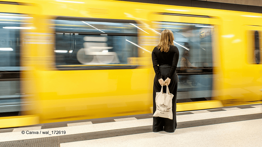 Eine Berliner U-Bahn fährt in einem Bahnhof ein. Eine Frau steht mit dem Rücken zu uns, sie trägt komplett schwarz und hält einen Jutebeutel hinter ihrem Rücken. Im Artikel geht es um einen Wegeunfall in einer U-Bahn.