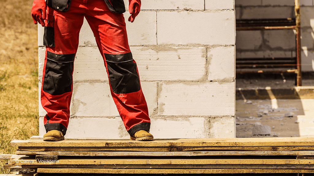 Eine Person in roter Arbeitskleidung steht auf einem Stapel Holzbretter. Es sind nur die Beine und Hände zusehen.