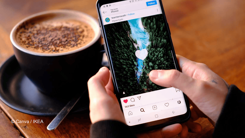 Zu sehen sind zwei Hände, die ein Handy halten. Auf dem Bildschirm ist ein Post auf Instagram zu sehen, den der User anscheinend geliked hat. Im Hintergrund ist eine schwarze Tasse mit Kakao oder Kaffee zu sehen.
