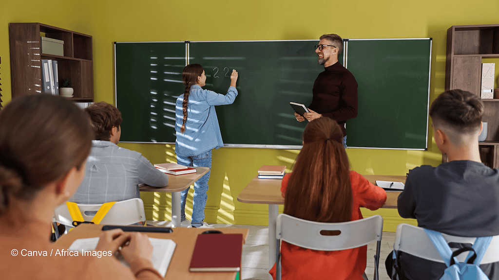Unterrichtsszene von einer Schulklasse mit Blick auf die Tafel. Vorne steht ein männlicher Lehrer an einer Kreidetafel. Daneben ein Mädchen, das etwas vorrechnet. Die anderen Schülerinnen und Schüler sind nur mit dem Rücken zu sehen. Symbolbild Probeunterricht