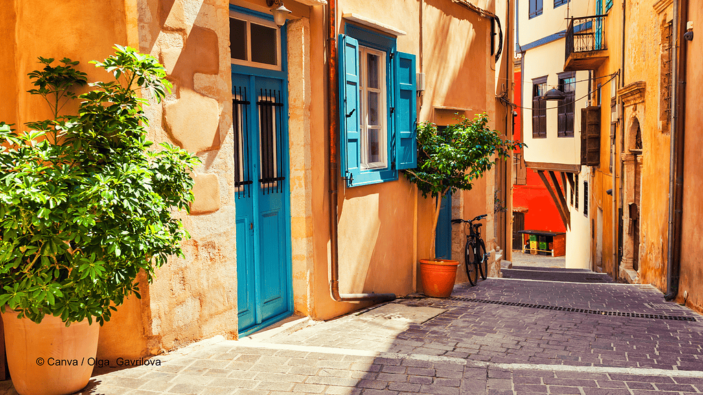 Ein Gasse in Chania auf Kreta, die Häuserfassaden sind orangegelb, Fensterläden und Türen strahlend blau. Am linken Rand steht eine große Topfpflanze.