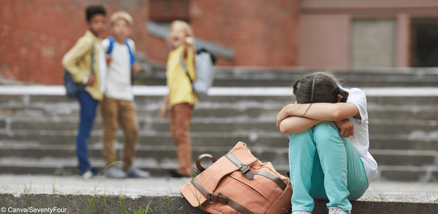 Kinder im Hintergrund zeigen auf ein Kind im Vordergrund, das sitzend den Kopf auf die Knie legt. Wie können Kinder vor Mobbing geschützt werden?