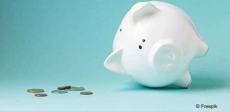Weißes Sparschwein, welches auf der Seite liegt, als wären die Finanzen in Schieflage geraten. Daneben ein paar Euromünzen vor hellblauem Hintergrund.