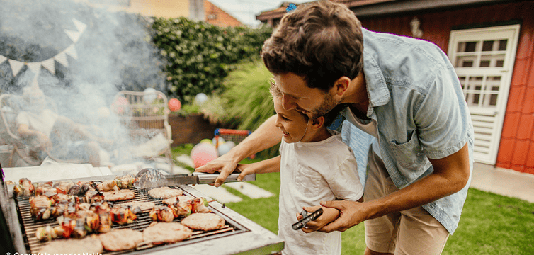Ein Junge und ein Mann grillen im Garten. Sie wenden einen Grillspieß mit einer Zange zusammen.