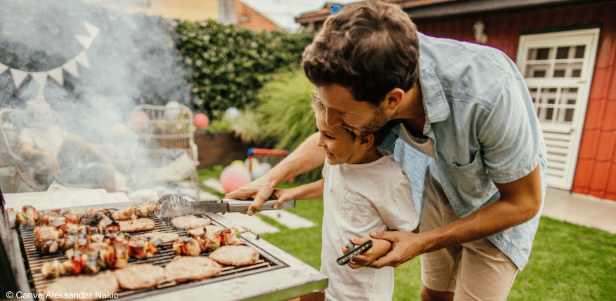 Ein Junge und ein Mann grillen im Garten. Sie wenden einen Grillspieß mit einer Zange zusammen.