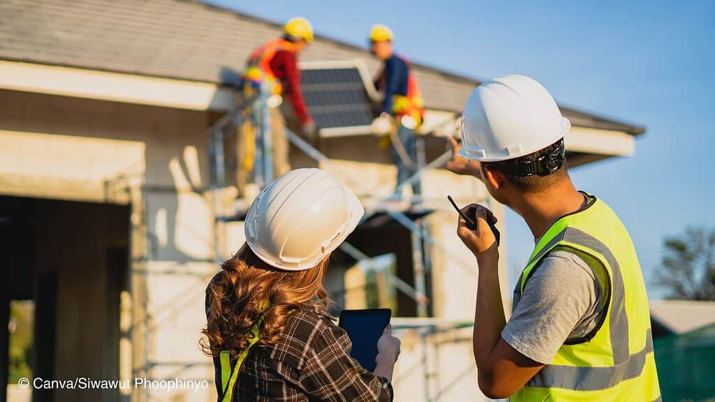 Im Vordergrund stehen eine Frau und ein Mann mit Schutzhelm und beobachten, wie zwei andere Menschen eine Photovoltaikanlage auf dem Dach eines neugebauten Hauses montieren.