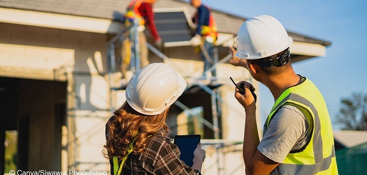 Im Vordergrund stehen eine Frau und ein Mann mit Schutzhelm und beobachten, wie zwei andere Menschen eine Photovoltaikanlage auf dem Dach eines neugebauten Hauses montieren.