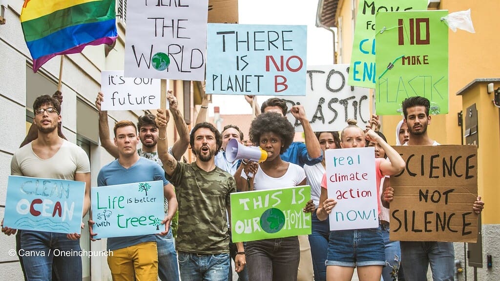 Eine Gruppe junger Menschen auf einer Demonstration. Mehrere junge Frauen und Männer halten Schilder mit umweltpolitischen Forderungen hoch. Eine Frau in der Mitte spricht in ein Megaphon. Links am Rand schwingt eine Person eine Regenbogenfahne.