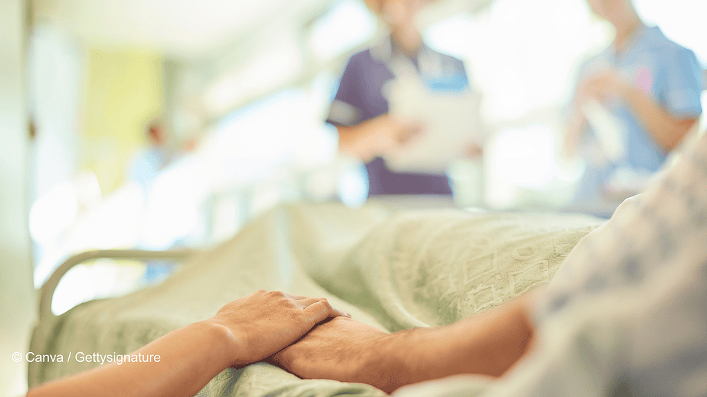 Szene im Krankenhaus in deren Vordergrund zwei Hände zu sehen sind. Auf der rechten Seite ist ein Krankenbett, die Person, die darin liegt hält die Hand einer Person auf der linken Seite. Beide sind nicht vollständig zu sehen. Im Hintergrund sind verschwommen zwei Menschen im Kasack. Symbolisch steht das Bild für einen Krankenhausaufenthalt nach einer Lipödem-Operation.