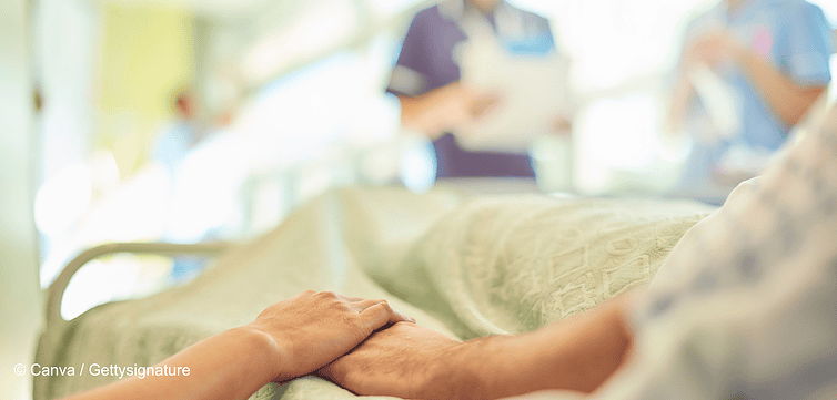 Szene im Krankenhaus in deren Vordergrund zwei Hände zu sehen sind. Auf der rechten Seite ist ein Krankenbett, die Person, die darin liegt hält die Hand einer Person auf der linken Seite. Beide sind nicht vollständig zu sehen. Im Hintergrund sind verschwommen zwei Menschen im Kasack. Symbolisch steht das Bild für einen Krankenhausaufenthalt nach einer Lipödem-Operation.